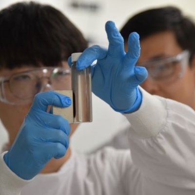 A close up of gloved hands holding a magnet against a small vial of brown liquid with two faces in the background